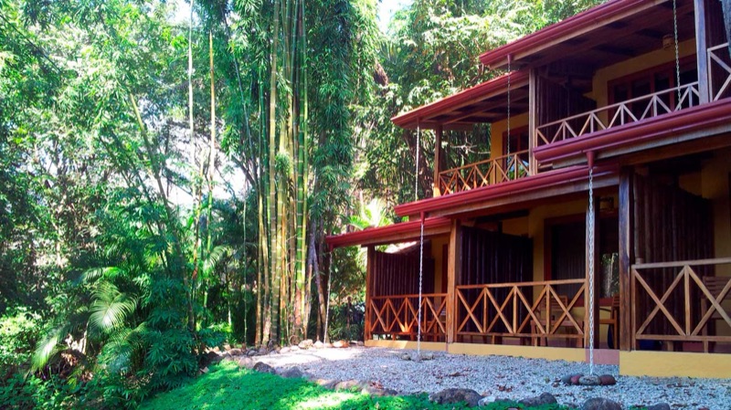 Two-story yellow building with red roofs and wooden balconies beside tall bamboo and garden at Esencia Hotel & Villas
